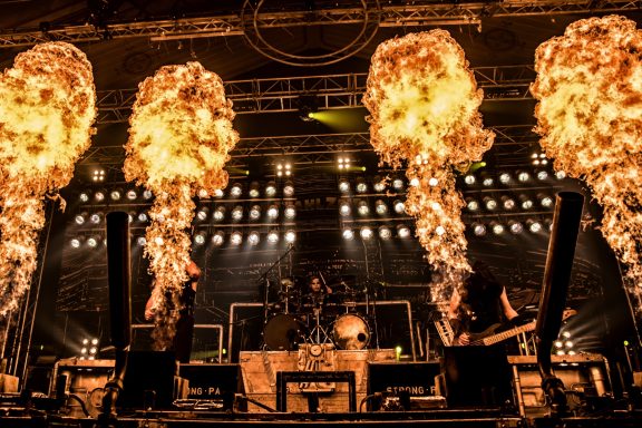 Feurige Effekte auf der Bühne bei einem Konzert mit Musikern im Hintergrund.