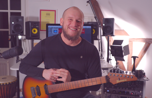 Lächelnder Musiker mit Gitarre in einem Studio voller Musikinstrumente.