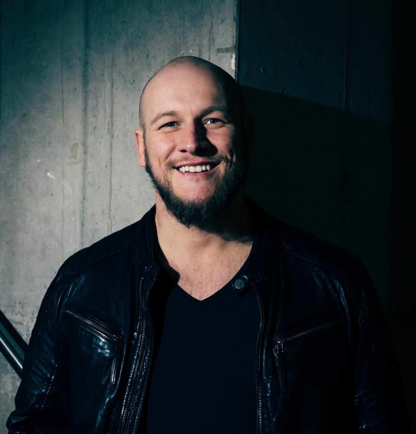Tobias Mertens Team Building Musik Lächelnder Mann mit Glatze trägt eine schwarze Lederjacke vor einer dunklen Wand.