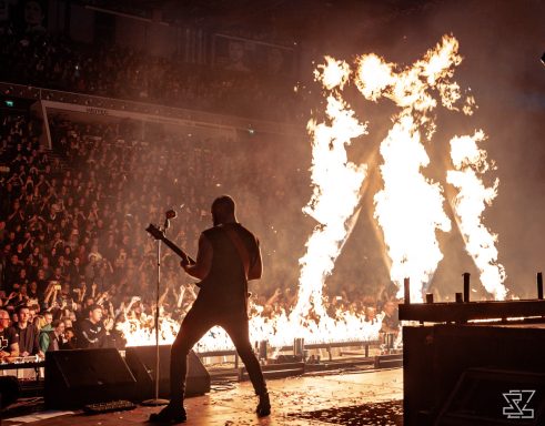 Musiker spielt Bass vor flammenden Pyroeffekten und begeistertem Publikum.